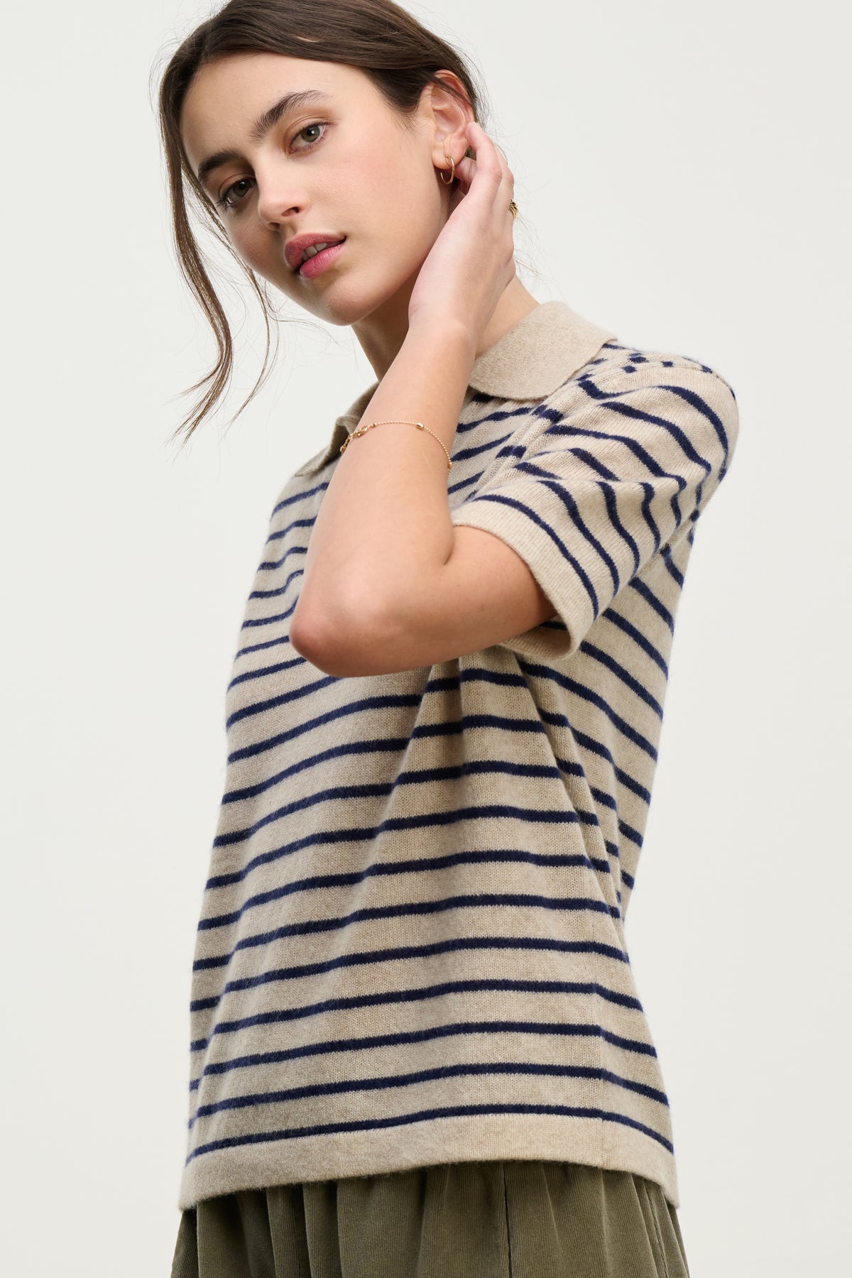   A woman wears the YAEL CASHMERE SWEATER POLO by Velvet by Graham & Spencer—a short-sleeved beige and navy knit top with stripes and a collar—standing against a plain light background. 