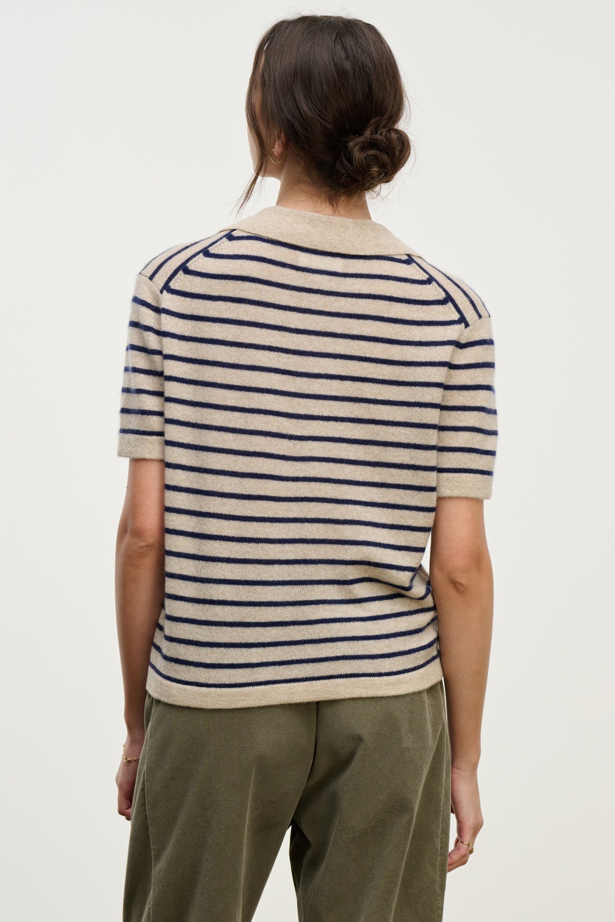  A person with long brown hair tied back is shown from the back wearing the YAEL CASHMERE SWEATER POLO by Velvet by Graham & Spencer, a short-sleeved, cropped sweater with beige and navy stripes, paired with green pants. 