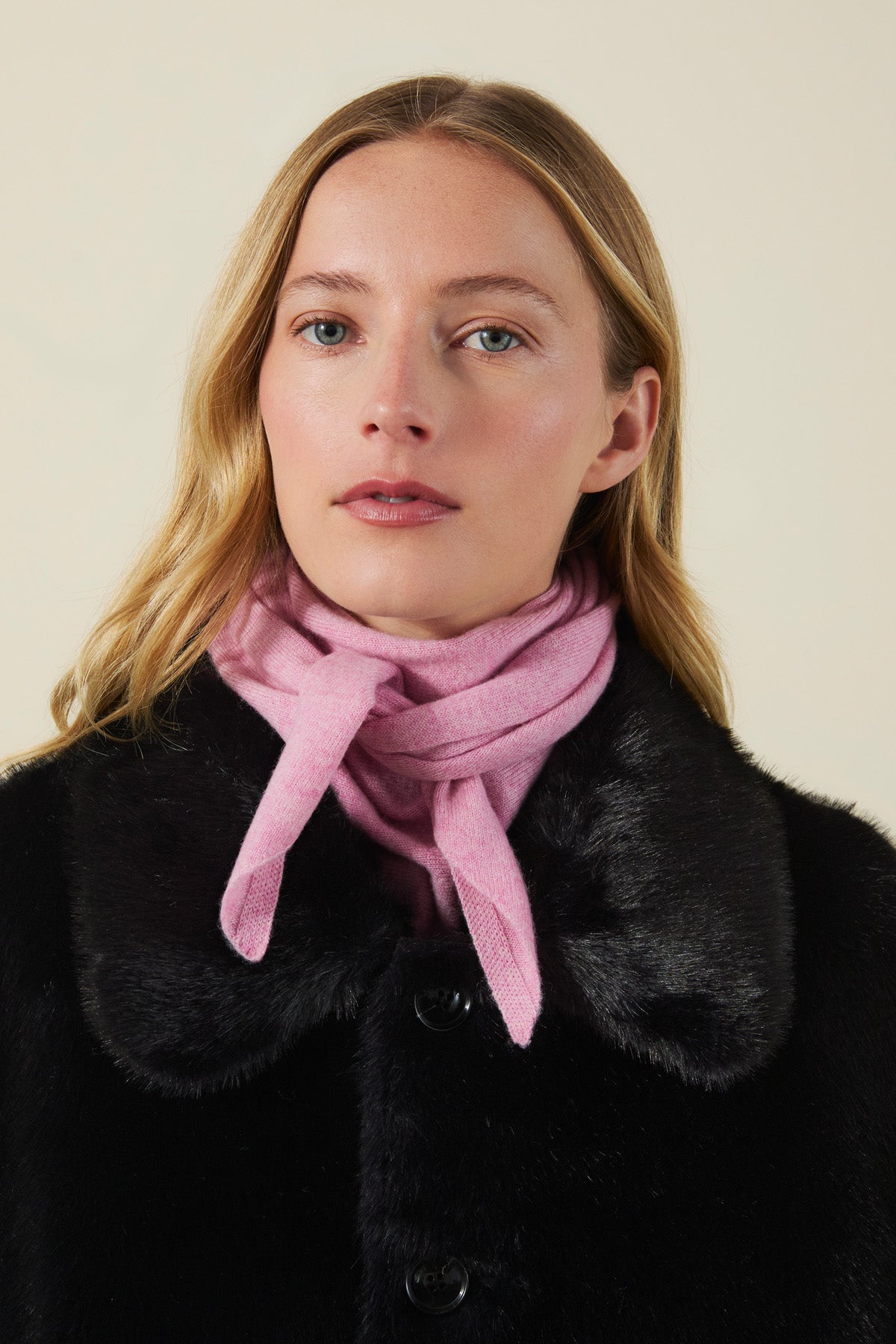 A person wearing a pink CLEO CASHMERE TRIANGLE SCARF knotted at the neck over a black collared coat.