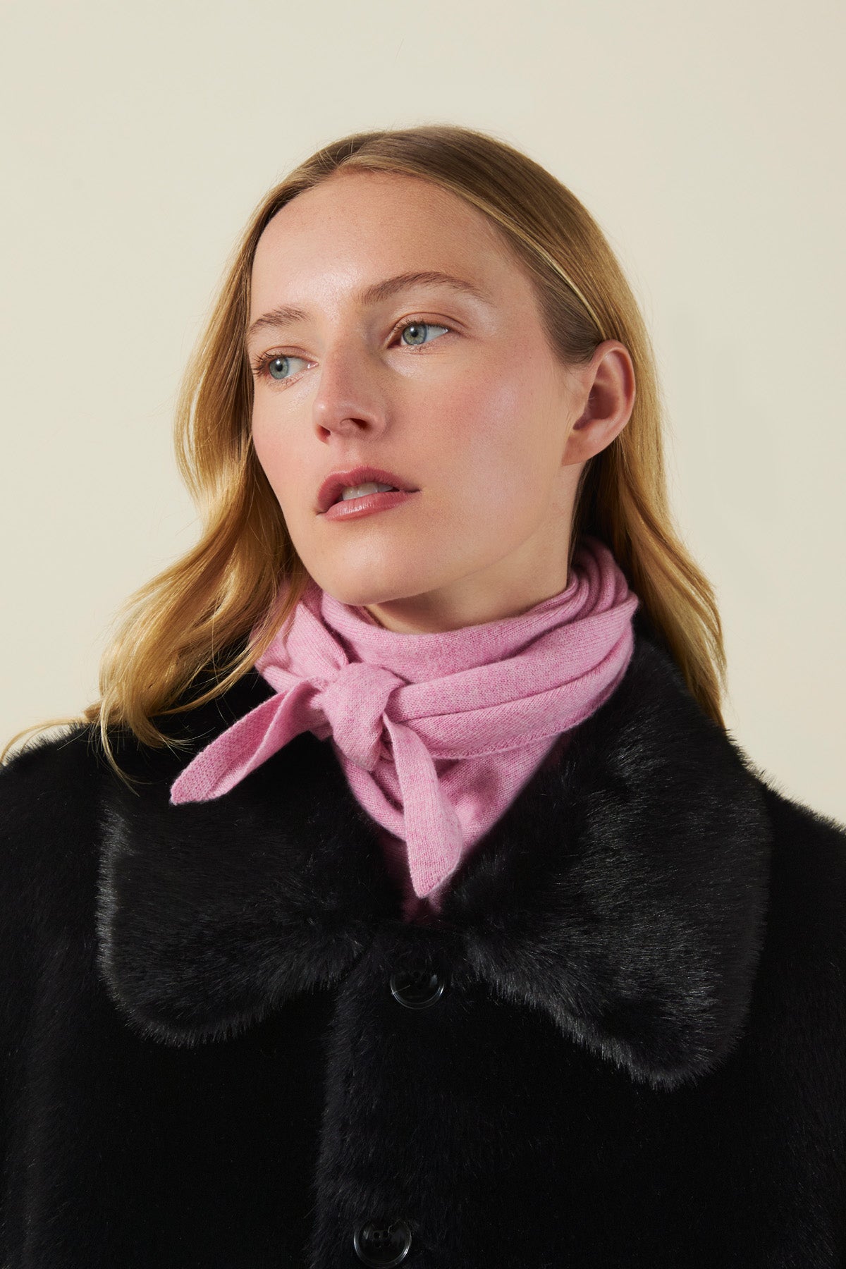 A person wears a small, pale pink CLEO CASHMERE TRIANGLE SCARF knotted around their neck under a black coat.-46490847019201
