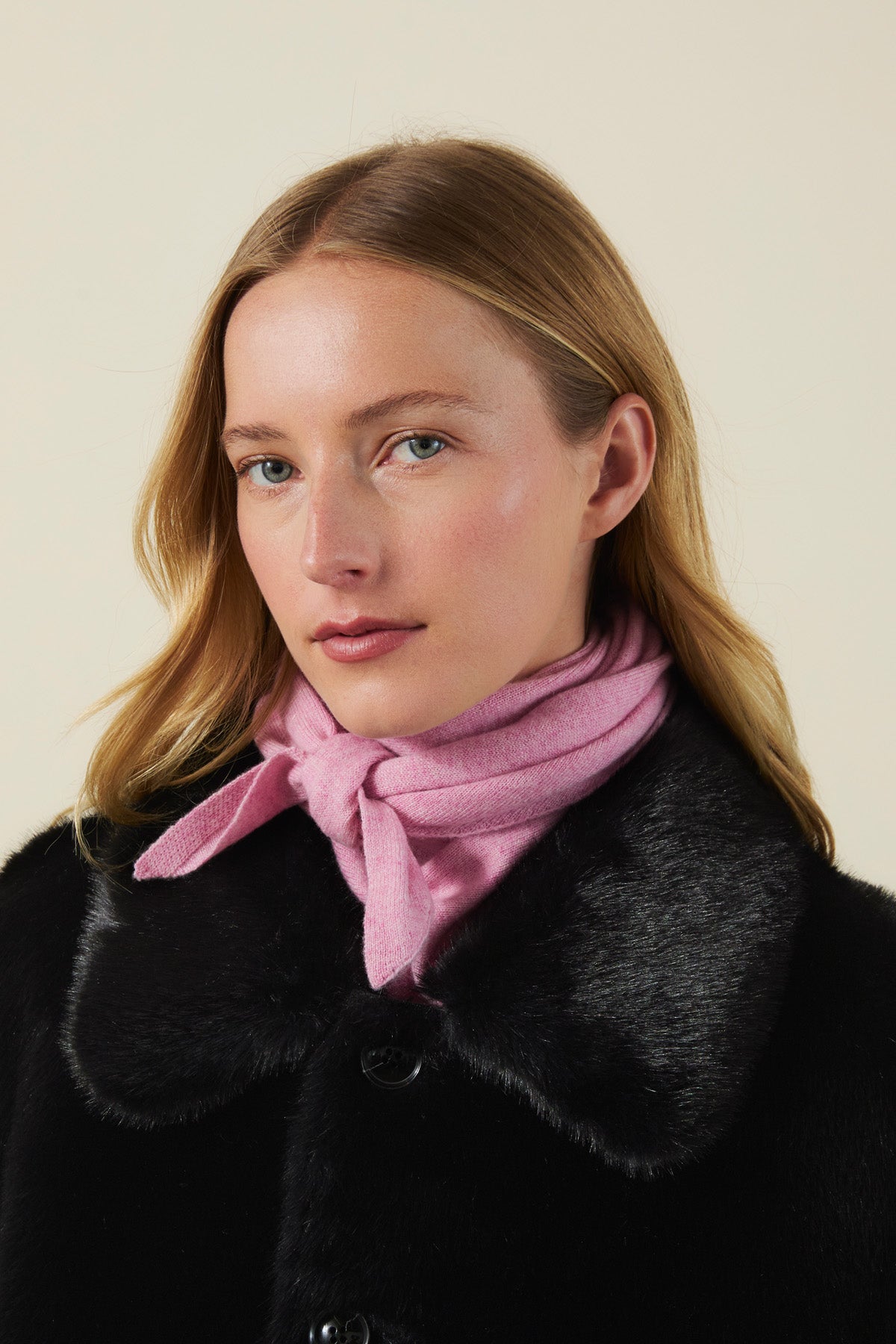 A person wearing a soft pink CLEO CASHMERE TRIANGLE SCARF tied at the neck under a black coat with a fur collar.