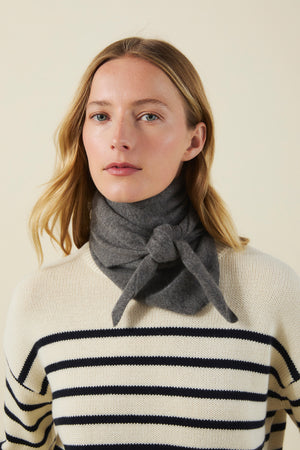 A woman wears a grey CLEO CASHMERE TRIANGLE SCARF tied around her neck over a cream and navy striped sweater.
