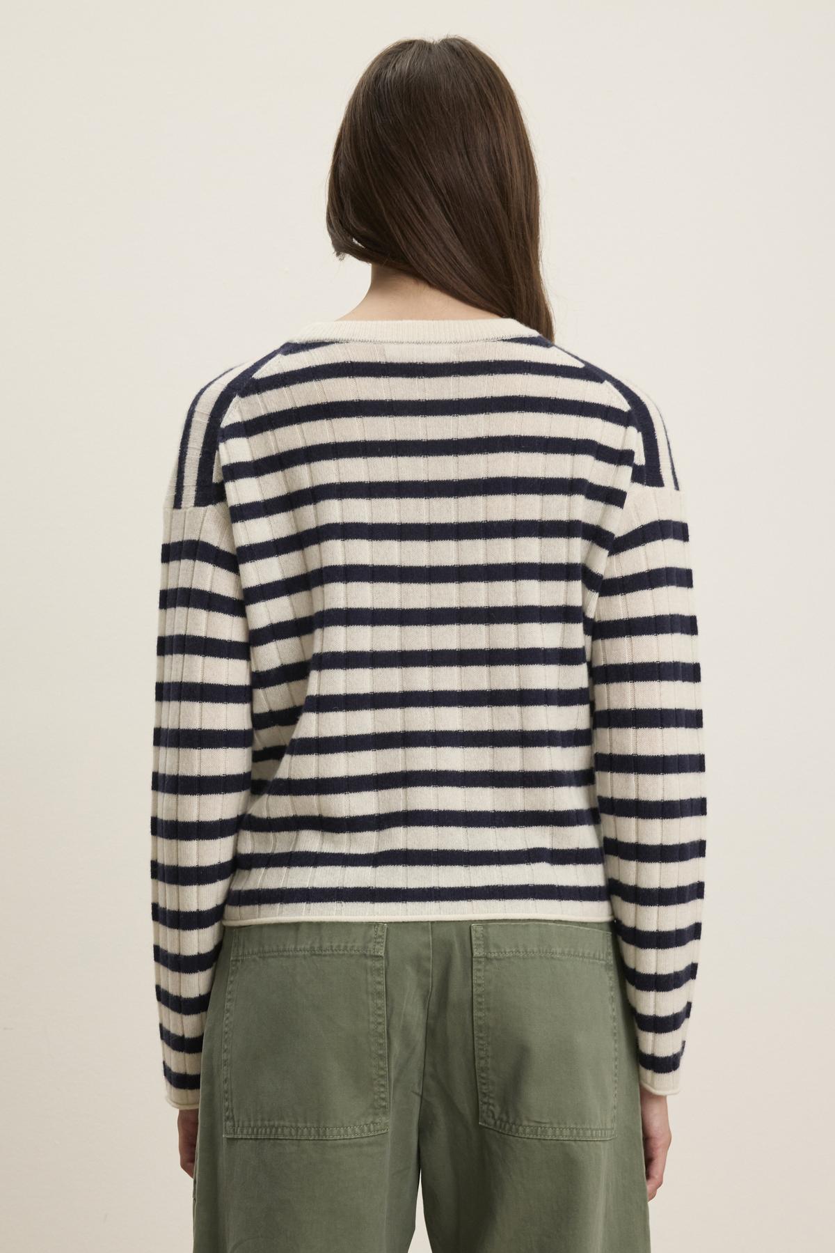   A person stands facing away, wearing the Velvet by Graham & Spencer ABI CASHMERE SWEATER in cream and black stripes with green pants against a plain background. 