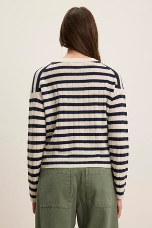 A person stands facing away, wearing the Velvet by Graham & Spencer ABI CASHMERE SWEATER in cream and black stripes with green pants against a plain background.