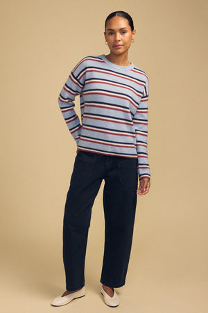 A person wearing the BETH SWEATER, a cozy knit crew neck with horizontal red, navy, and white stripes, paired with dark trousers and light-colored slip-on shoes.