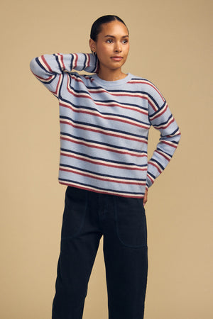 A person wearing a BETH SWEATER, featuring a cozy knit crew neck with horizontal red and navy stripes, paired with dark pants.