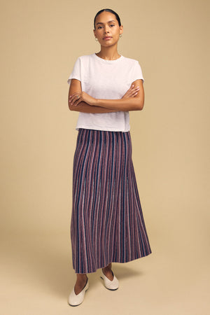 A person wearing a white short-sleeved t-shirt, a DESIREE FLARED SKIRT with vertical blue and red stripes, and cream-colored slip-on shoes.