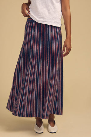 A person wears a mid-length, flared DESIREE FLARED SKIRT by Velvet by Graham & Spencer featuring vertical multicolored stripes, paired with a white t-shirt and white pointed-toe shoes.