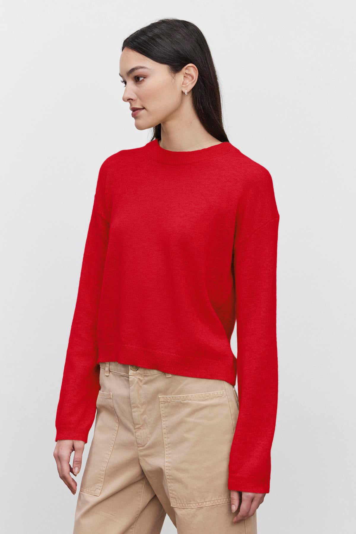   A person wearing a BROOKLYN CASHMERE SWEATER by Velvet by Graham & Spencer, featuring a crew neckline in red, paired with beige pants is standing against a plain background, looking to the side. 