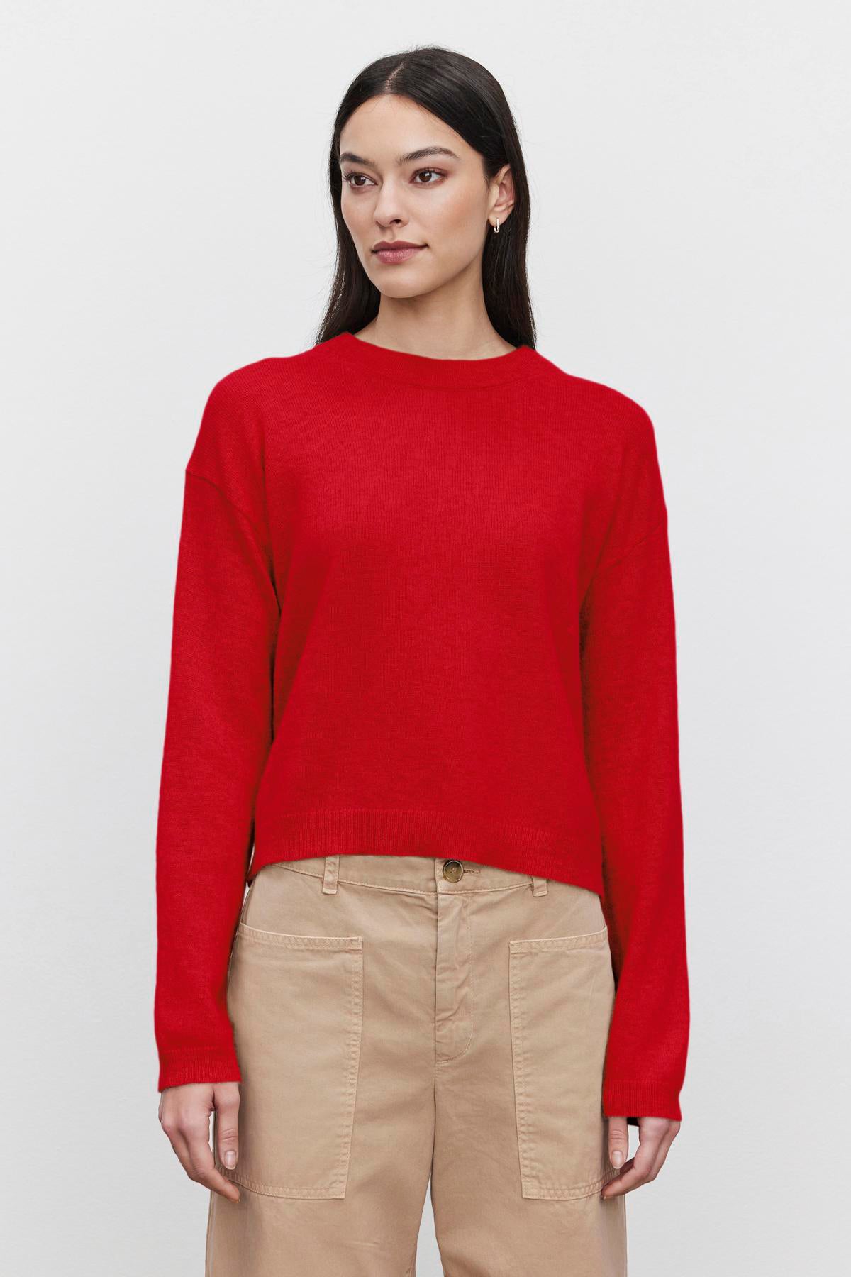   A woman with long dark hair wears the BROOKLYN CASHMERE SWEATER from Velvet by Graham & Spencer, featuring a plain red design and a crew neckline, paired with beige pants, as she stands against a white background. 