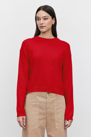 A woman with long dark hair wears the BROOKLYN CASHMERE SWEATER from Velvet by Graham & Spencer, featuring a plain red design and a crew neckline, paired with beige pants, as she stands against a white background.