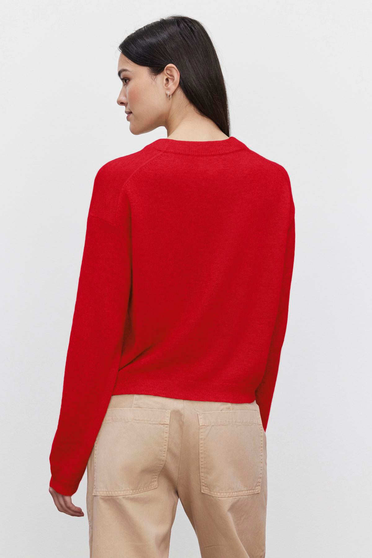   A person with long dark hair is facing away, wearing the Velvet by Graham & Spencer BROOKLYN CASHMERE SWEATER in red and beige pants, showcasing a contemporary look against a white background. 