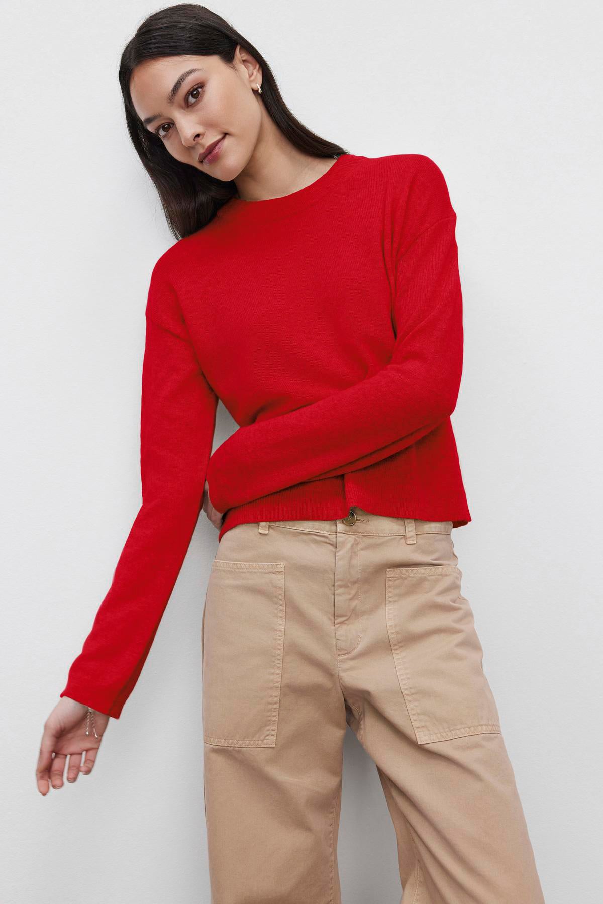   A woman wearing a BROOKLYN CASHMERE SWEATER by Velvet by Graham & Spencer in red, paired with beige pants, stands against a plain white background. 