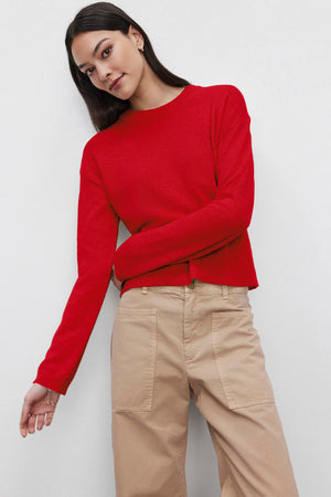 A woman wearing a BROOKLYN CASHMERE SWEATER by Velvet by Graham & Spencer in red, paired with beige pants, stands against a plain white background.