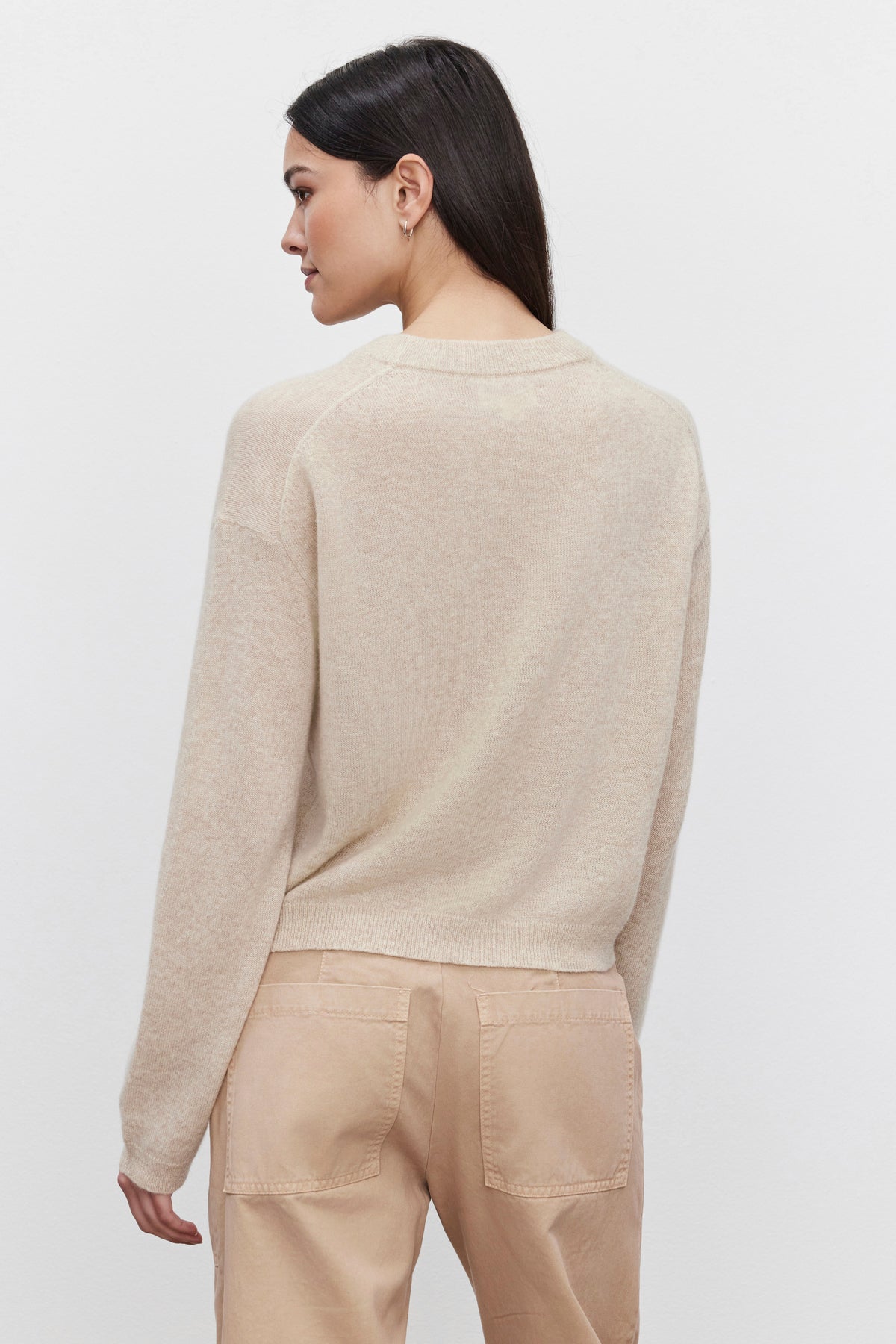   A person with long black hair is shown from the side and back, wearing the Velvet by Graham & Spencer BROOKLYN CASHMERE SWEATER in beige, paired with matching beige pants, capturing a contemporary look. 