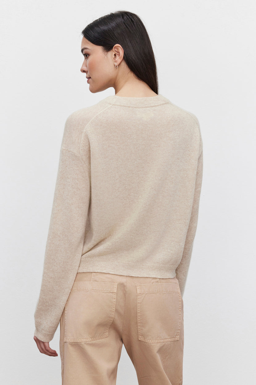 A person with long black hair is shown from the side and back, wearing the Velvet by Graham & Spencer BROOKLYN CASHMERE SWEATER in beige, paired with matching beige pants, capturing a contemporary look.