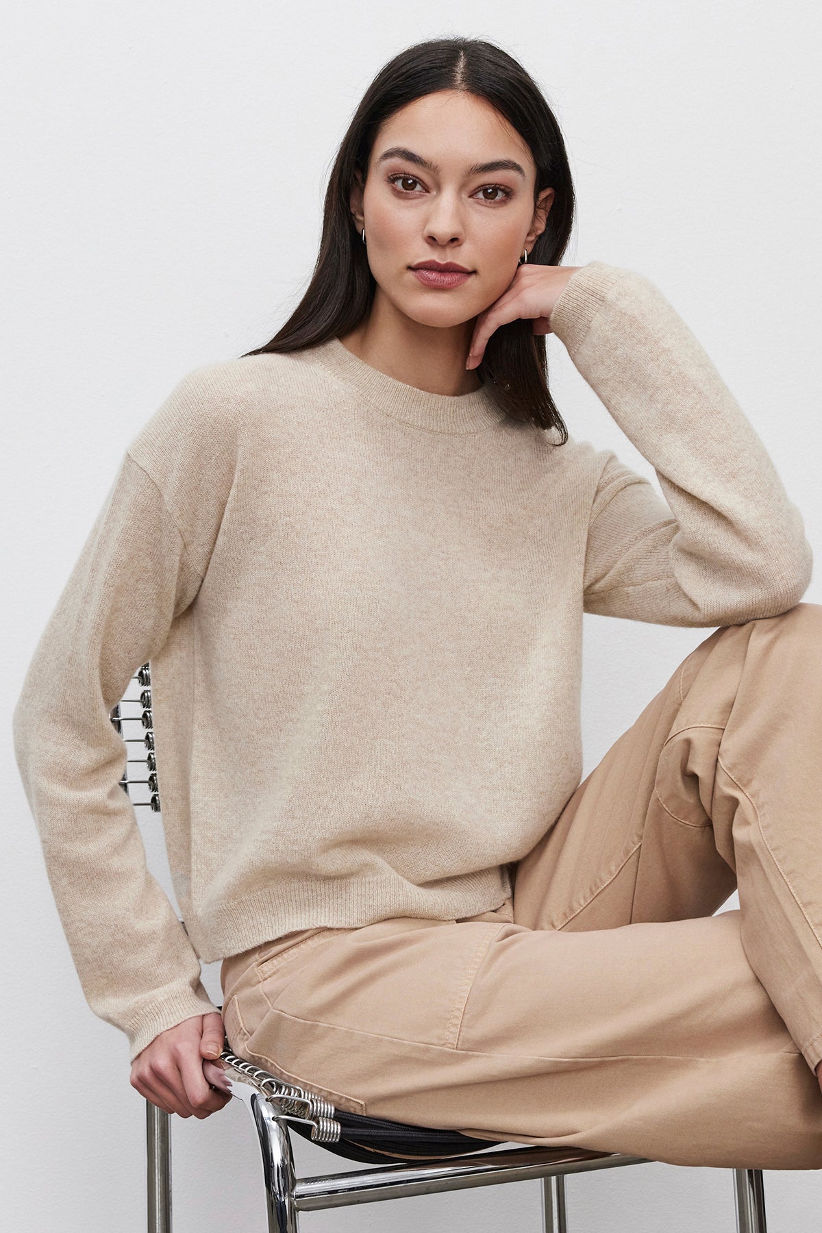   A person with long hair wearing a BROOKLYN CASHMERE SWEATER by Velvet by Graham & Spencer and khaki pants sits on a metal chair, resting their chin on their hand against a plain white background. 