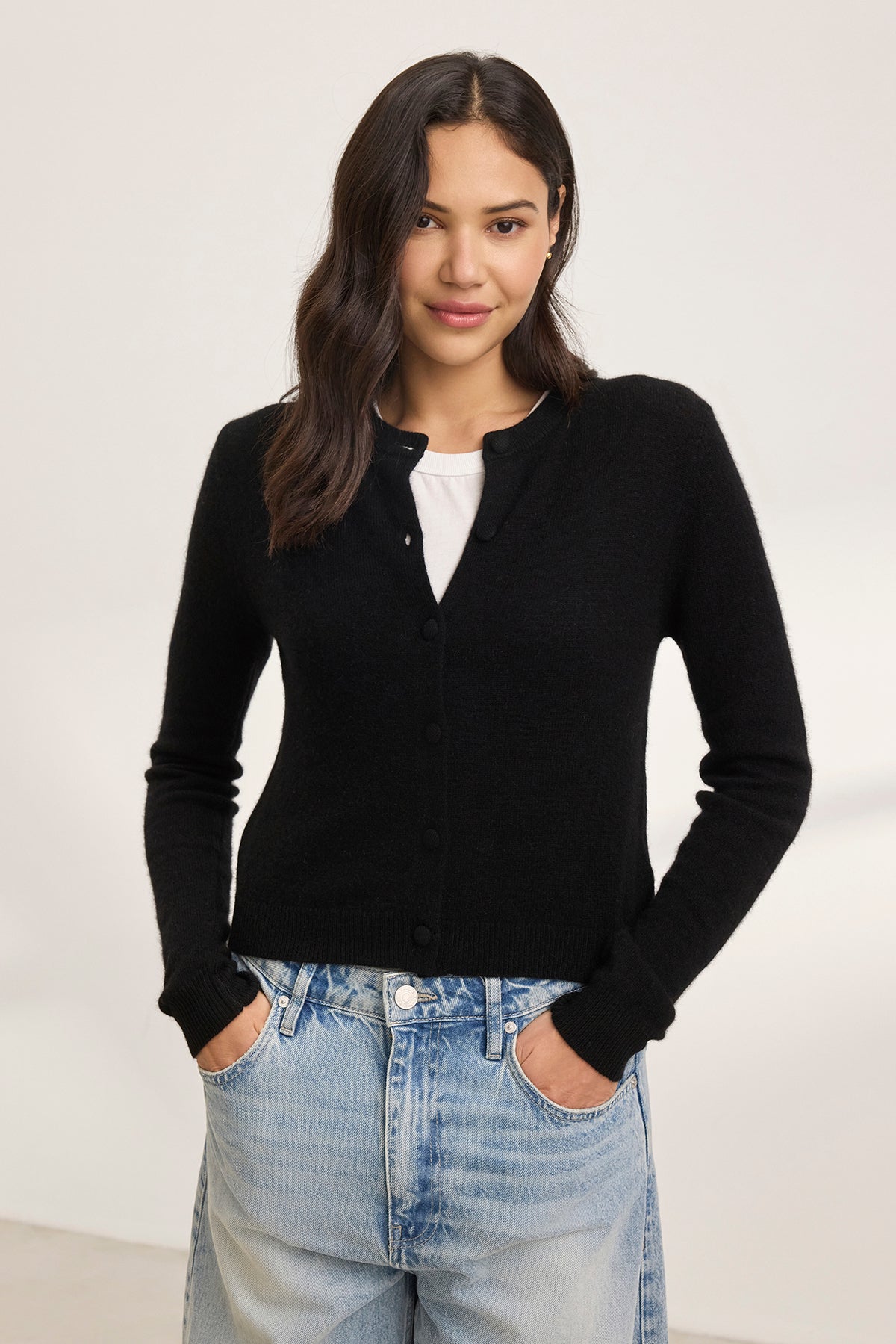 A woman stands against a plain background, wearing the CHRICHET CASHMERE CARDIGAN from Velvet by Graham & Spencer over a white shirt, paired with blue jeans.-38754141307073