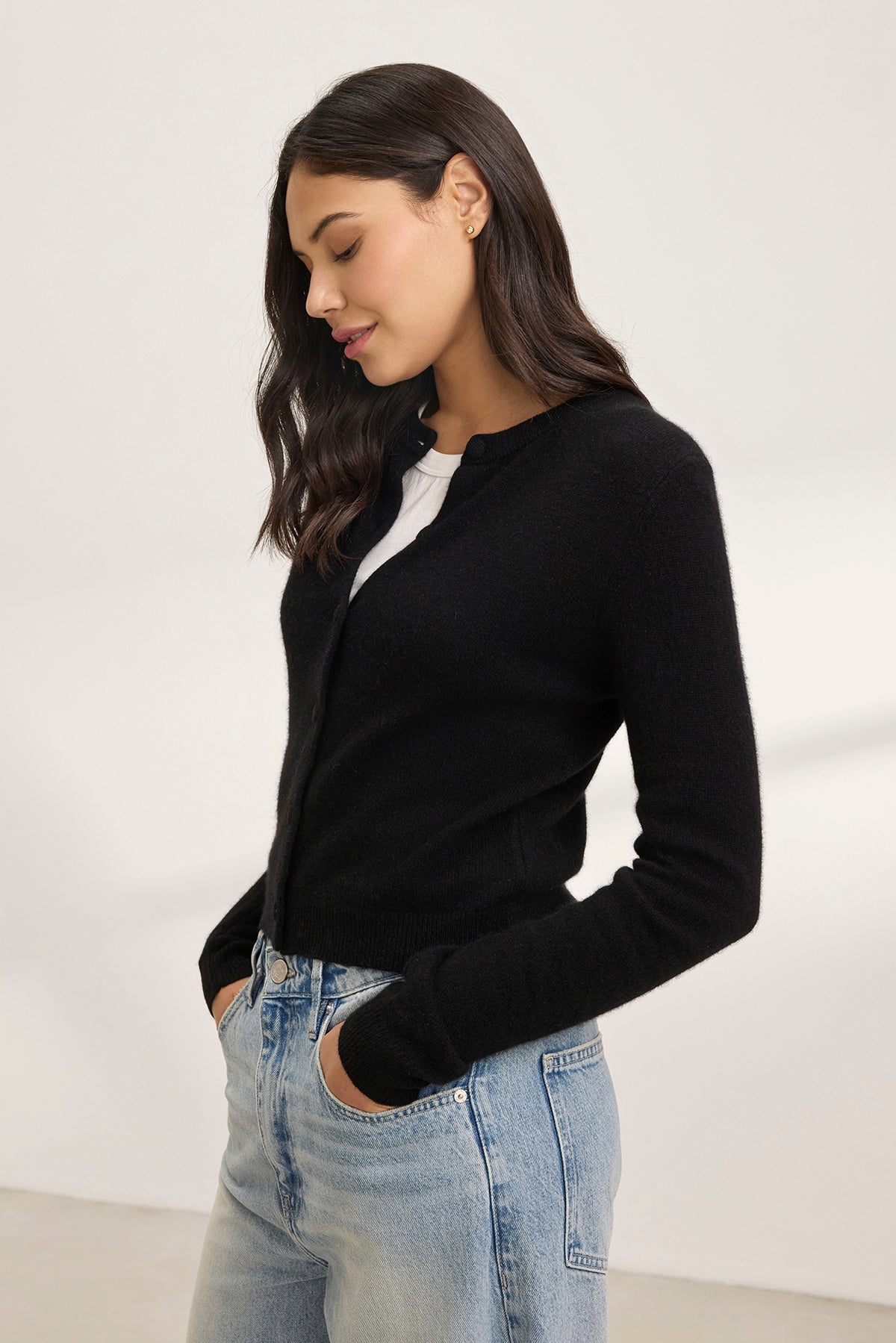 A woman wearing the CHRICHET CASHMERE CARDIGAN by Velvet by Graham & Spencer and blue jeans stands with hands in pockets against a plain background.-38754141339841