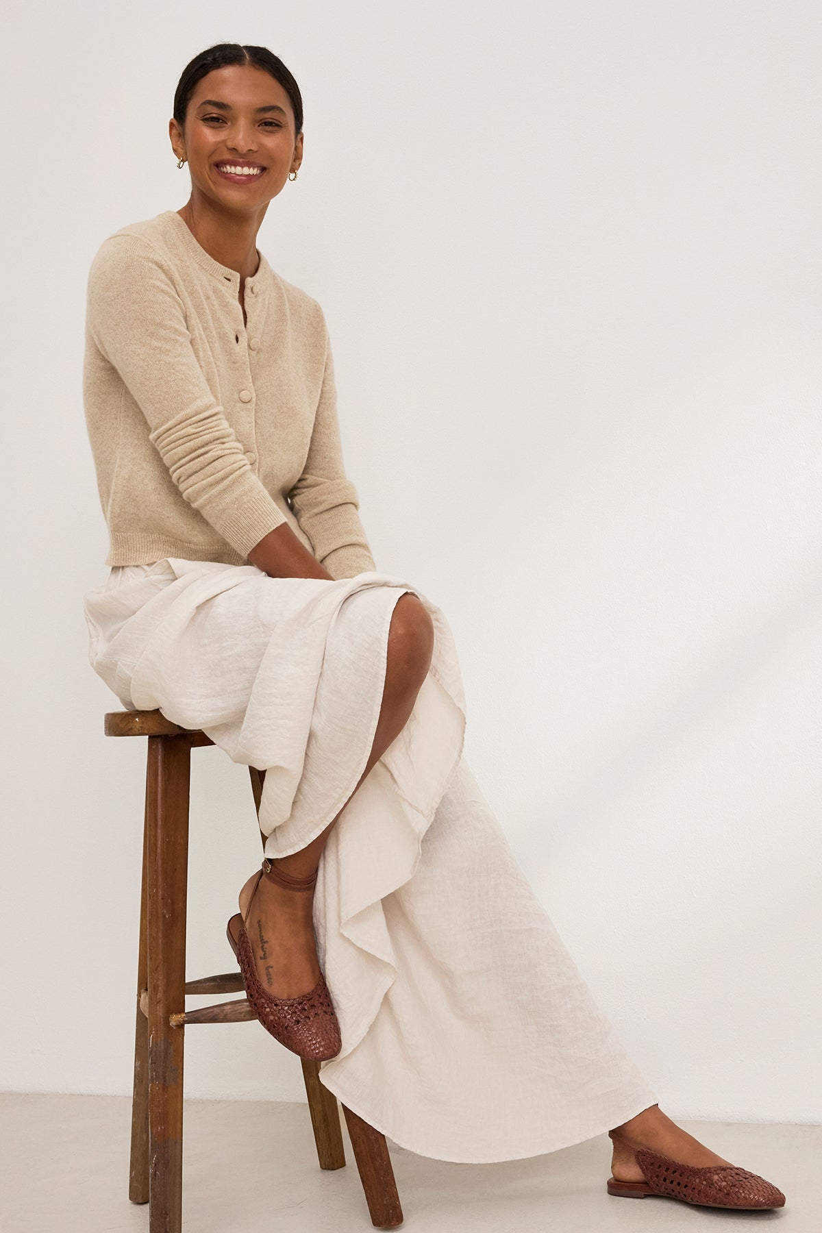 A person on a stool smiles against a plain background, wearing Velvet by Graham & Spencer's CHRICHET CASHMERE CARDIGAN in beige over a white skirt, paired with button-front brown woven shoes.-38850825027777