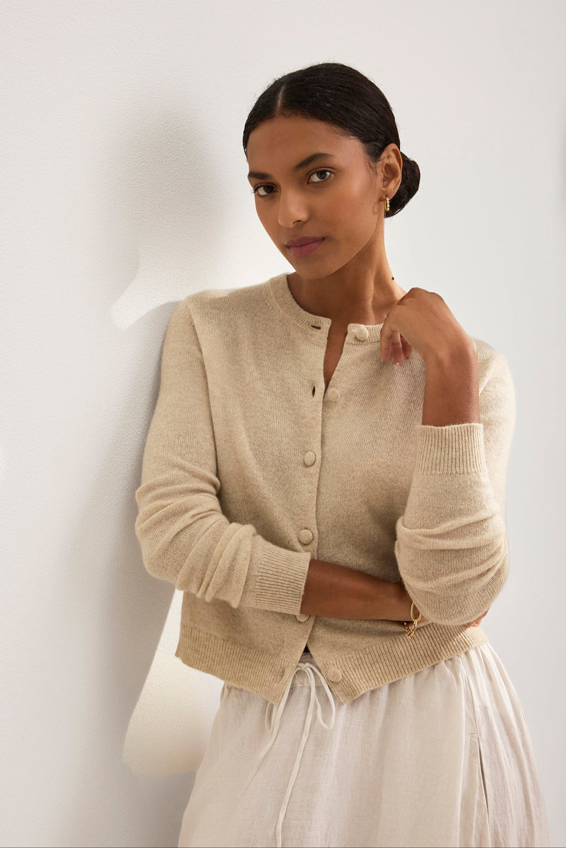 A person with dark hair in a low bun wears the CHRICHET CASHMERE CARDIGAN by Velvet by Graham & Spencer and a white skirt. They stand against a light-colored wall, gazing forward with one hand touching their neck.-38754141667521