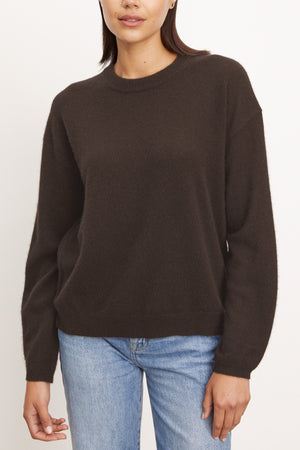 A person wearing the Velvet by Graham & Spencer BRYNNE CASHMERE SWEATER in dark brown with relaxed shoulders and light blue jeans stands against a plain, off-white background.