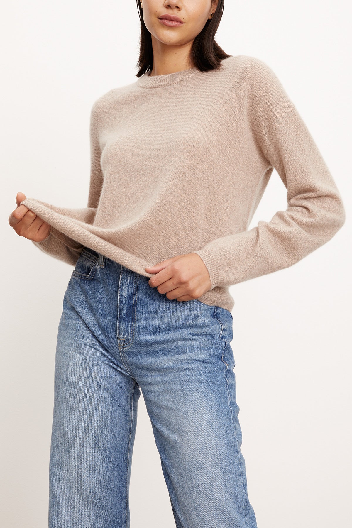 A woman wears the BRYNNE CASHMERE SWEATER by Velvet by Graham & Spencer—a beige crewneck with ribbed trims—paired with high-waisted blue jeans, holding out the hem of her sweater.-40278713565377