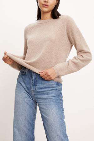 A woman wears the BRYNNE CASHMERE SWEATER by Velvet by Graham & Spencer—a beige crewneck with ribbed trims—paired with high-waisted blue jeans, holding out the hem of her sweater.