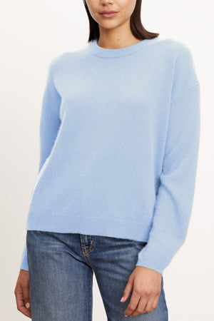 A person wearing the Velvet by Graham & Spencer BRYNNE CASHMERE SWEATER in light blue with ribbed trims and blue jeans, standing against a plain light background.
