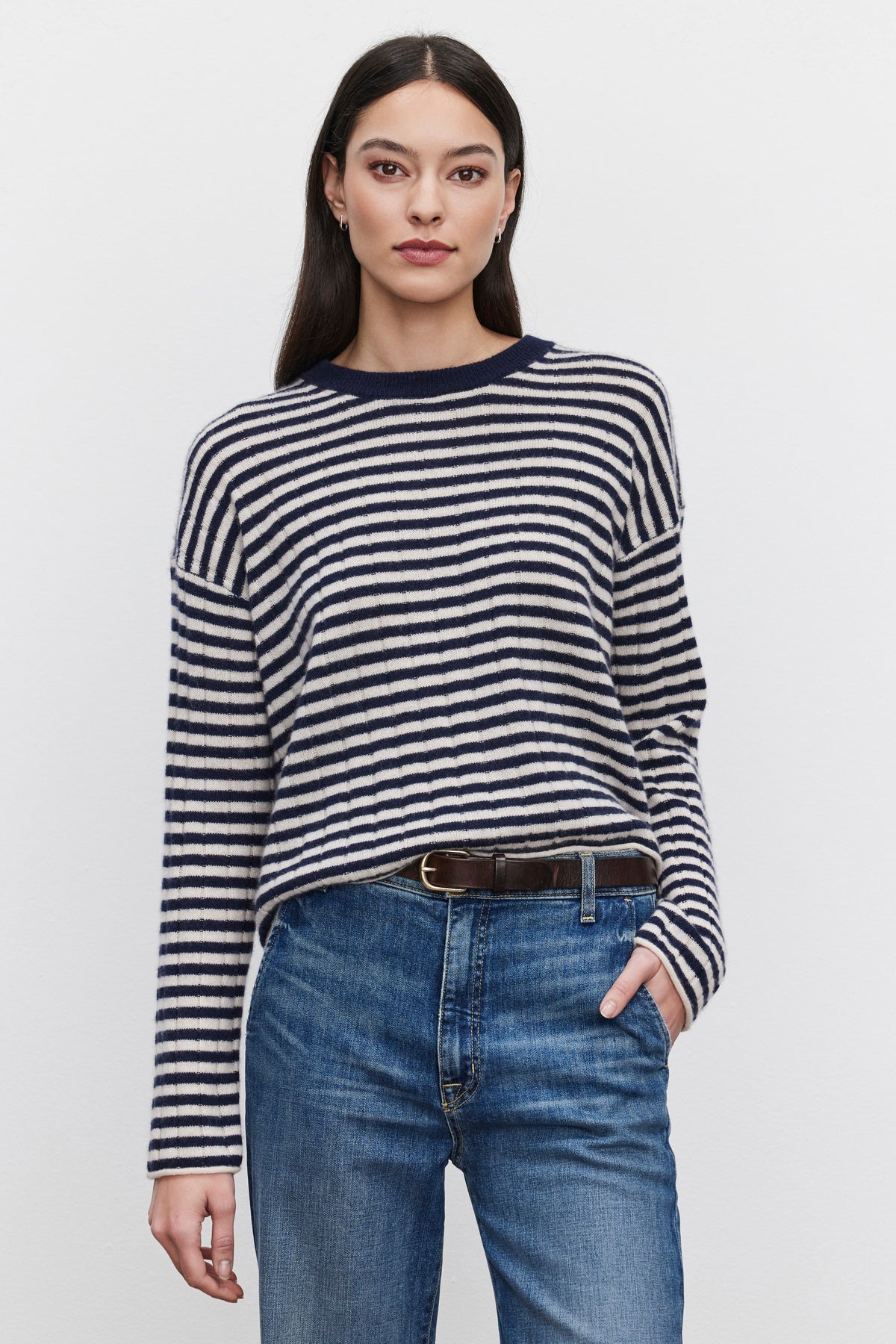   Person standing against a plain background, wearing a black and white striped Monica Cashmere Sweater by Velvet by Graham & Spencer, blue jeans, and a black belt, with one hand in their pocket. 