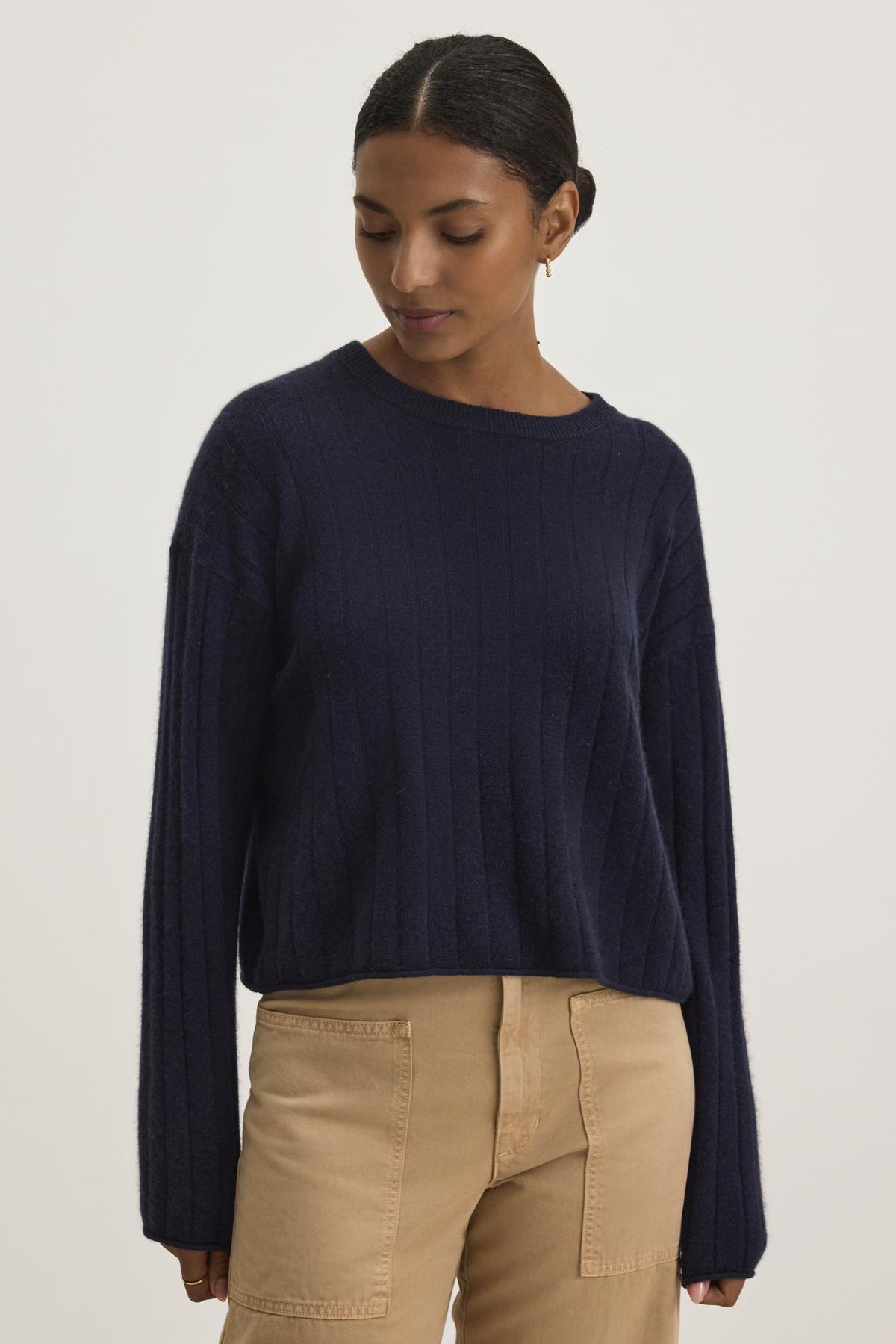   A person wearing the Monica Cashmere sweater in dark blue by Velvet by Graham & Spencer, paired with beige pants, looking down against a plain background. 