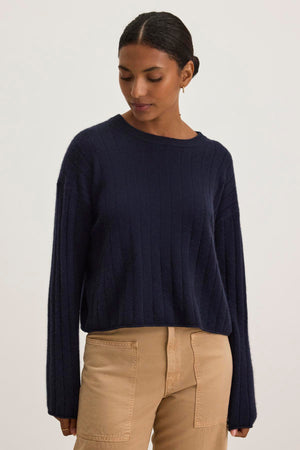 A person wearing the Monica Cashmere sweater in dark blue by Velvet by Graham & Spencer, paired with beige pants, looking down against a plain background.