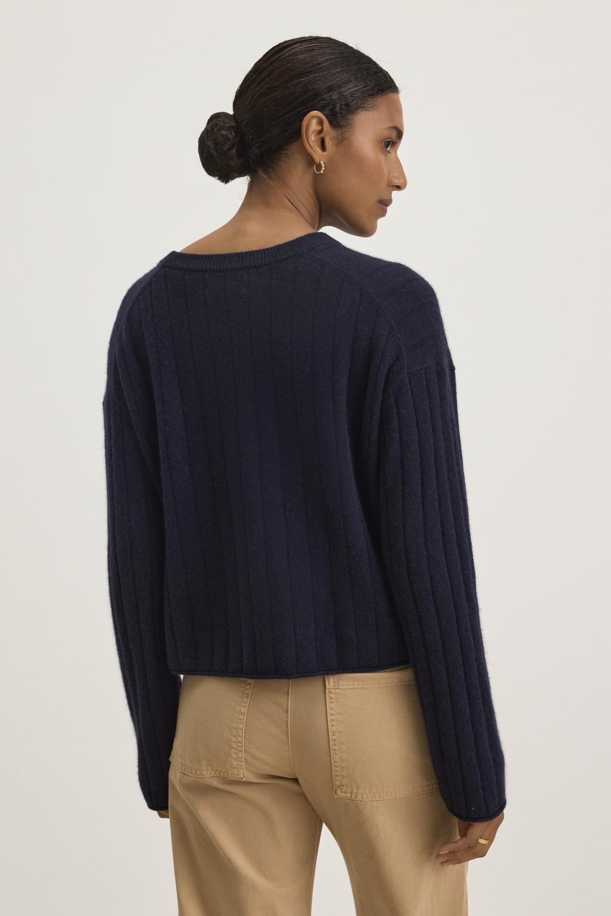   A person stands with their back to the camera, wearing the luxurious MONICA CASHMERE SWEATER by Velvet by Graham & Spencer, paired with beige pants, against a neutral background. 