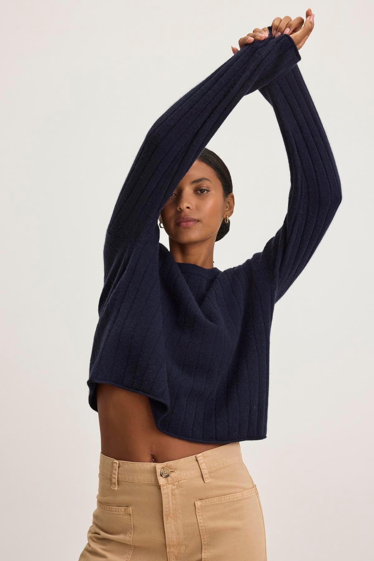   A person wearing the MONICA CASHMERE SWEATER by Velvet by Graham & Spencer in navy and beige pants, standing with arms raised against a plain background. 