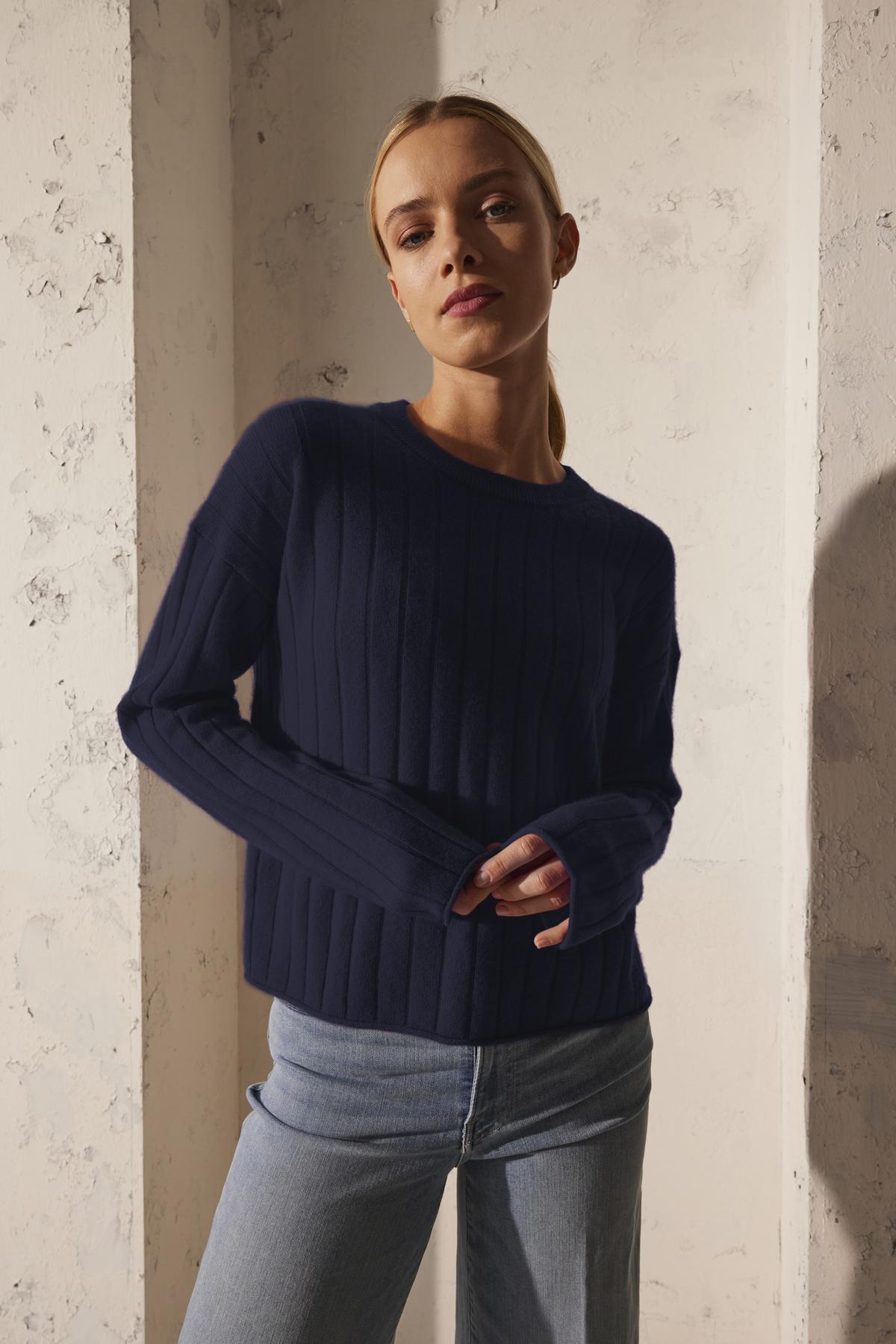   A person wearing the MONICA CASHMERE SWEATER by Velvet by Graham & Spencer and jeans stands against a textured wall in soft lighting, exuding luxurious comfort. 