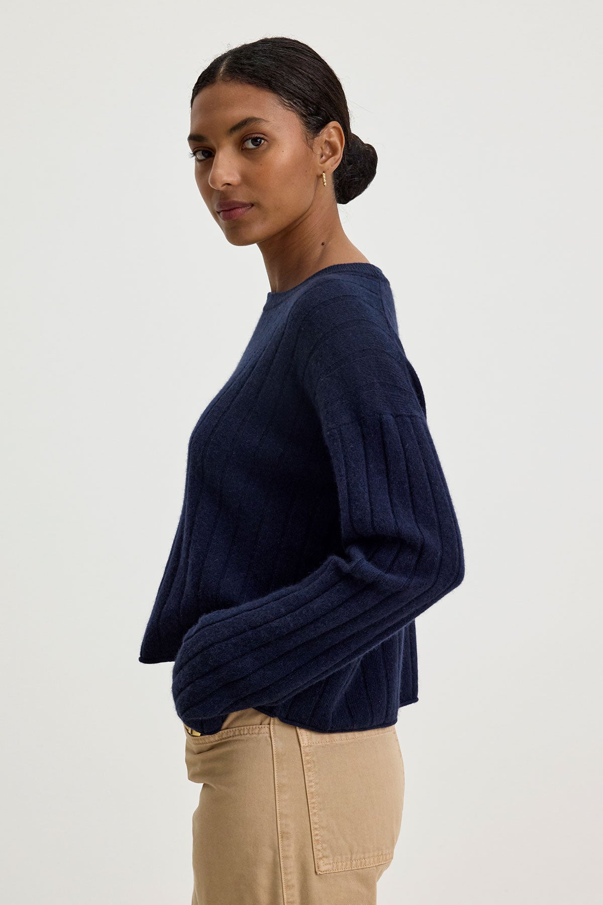   A woman with dark hair tied back, wearing a navy blue long-sleeve MONICA CASHMERE SWEATER by Velvet by Graham & Spencer and beige pants, stands against a plain background. 