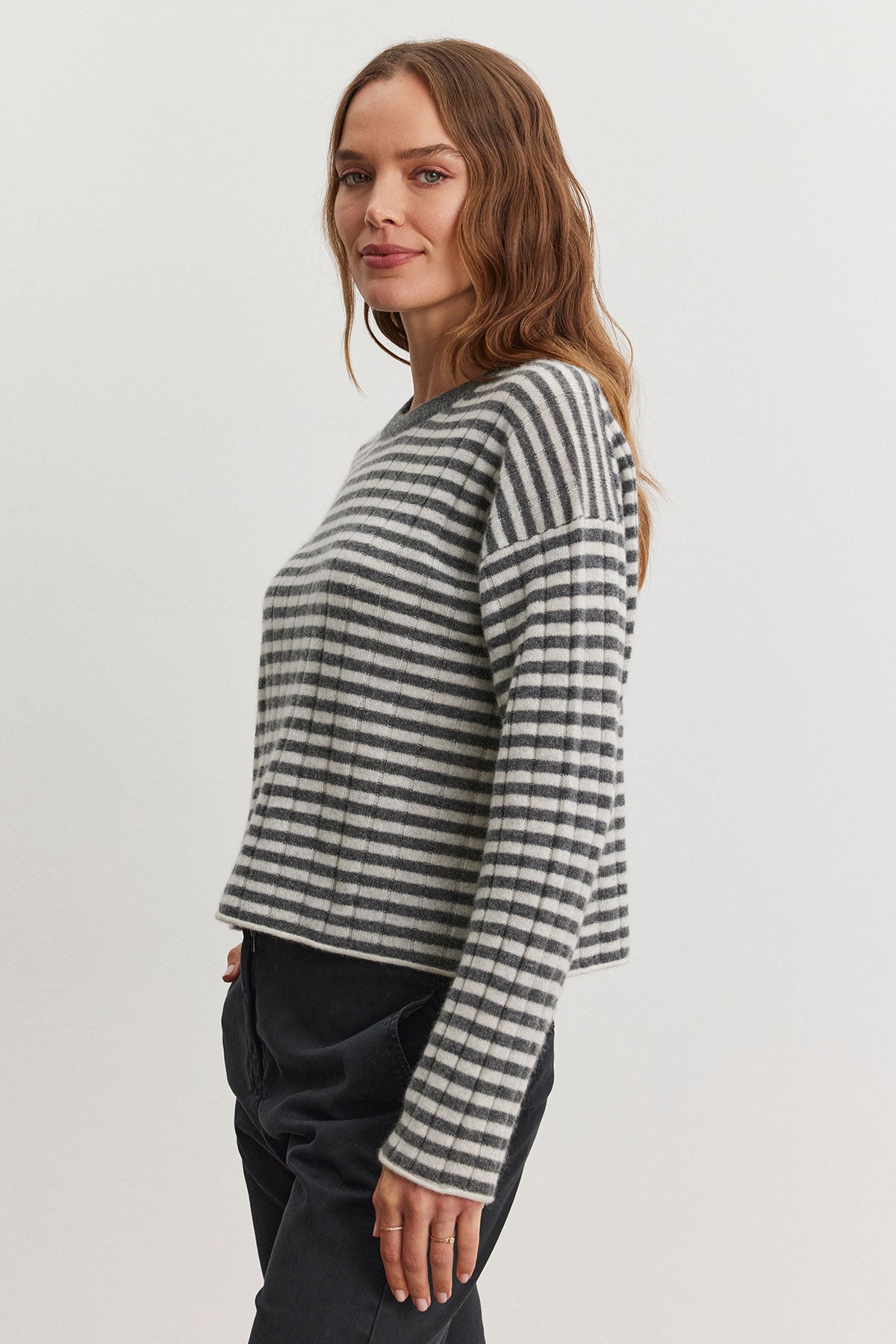 A woman wearing the MONICA CASHMERE SWEATER by Velvet by Graham & Spencer, featuring gray and white stripes, paired with dark pants, stands against a plain white background.-38301405282497