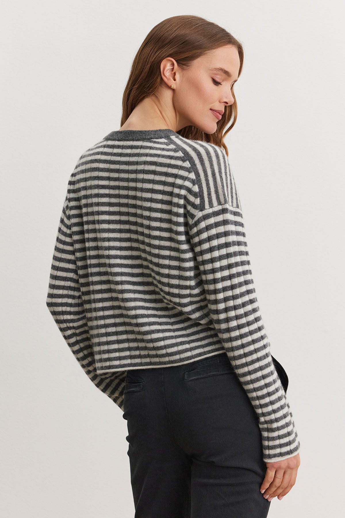   A person wearing the MONICA CASHMERE SWEATER by Velvet by Graham & Spencer, a wardrobe essential for luxurious comfort in black and white stripes, along with black pants stands against a plain background, facing slightly away. 