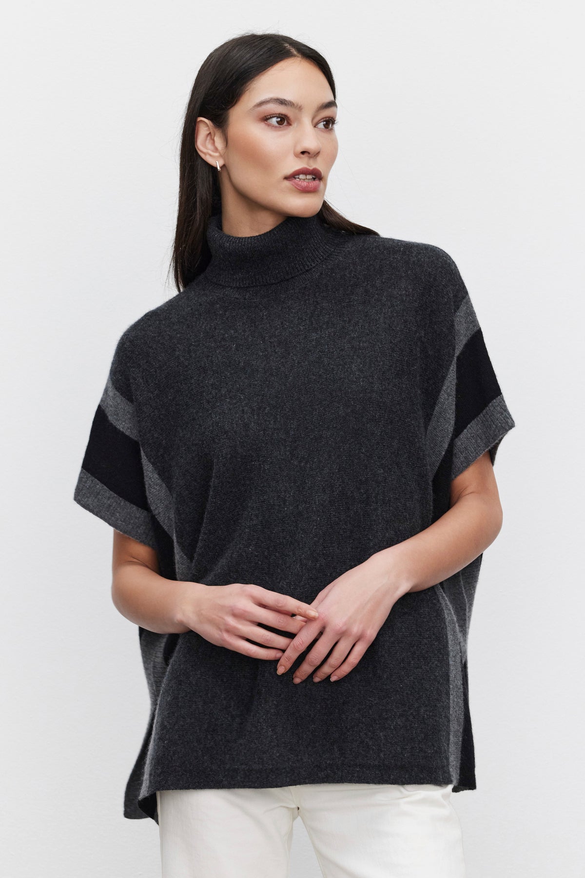   A woman with long dark hair wears the REESE CASHMERE TURTLENECK PONCHO by Velvet by Graham & Spencer and white pants, standing against a plain white background. 