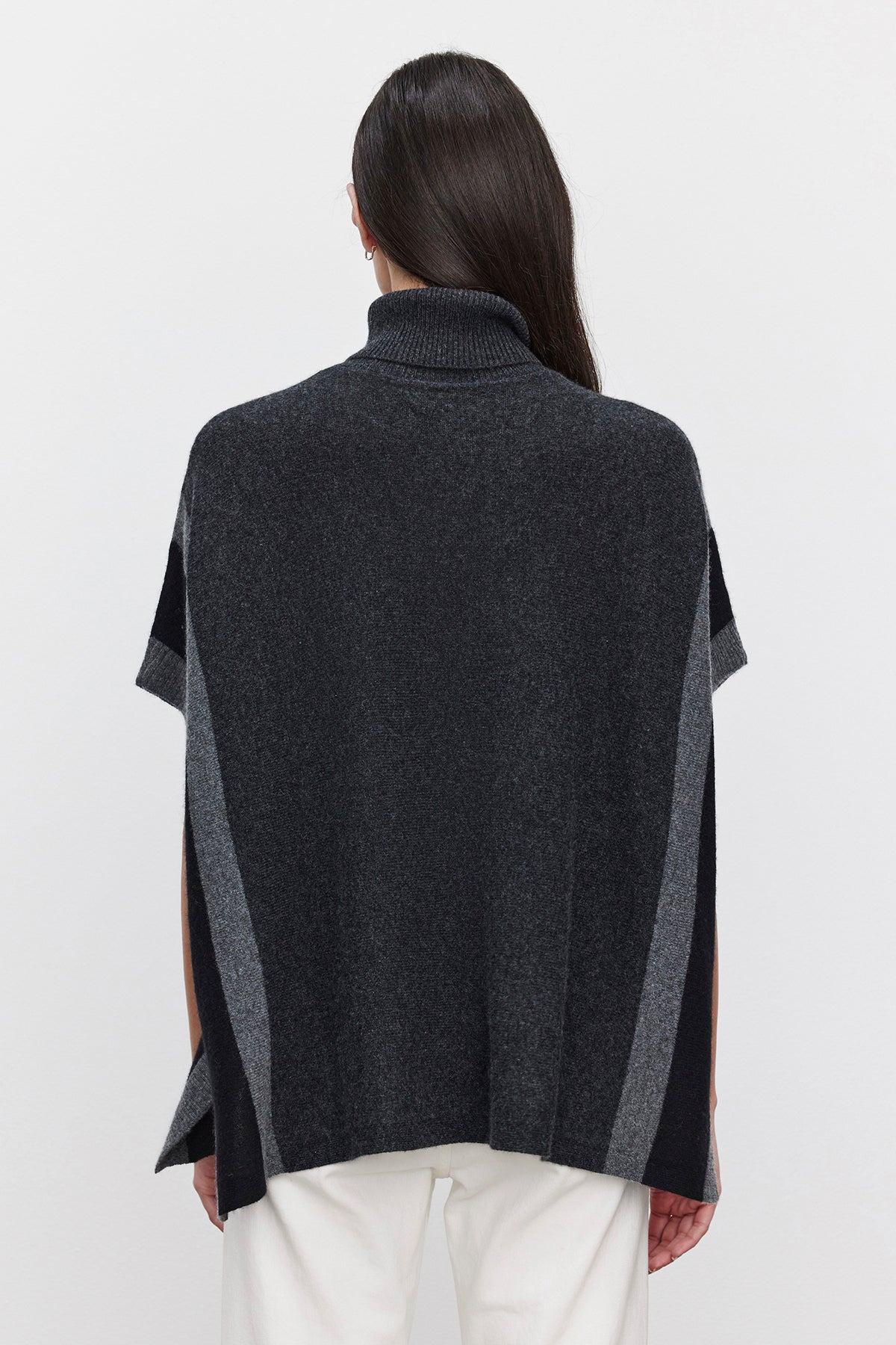   A person with long dark hair is wearing the REESE CASHMERE TURTLENECK PONCHO from Velvet by Graham & Spencer, featuring a dark gray hue with lighter gray vertical stripes and a turtleneck, facing away from the camera against a plain white background. The relaxed silhouette adds to the casual elegance of the outfit. 