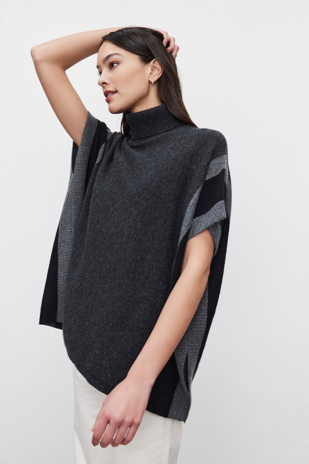   A person with long dark hair wears the REESE CASHMERE TURTLENECK PONCHO by Velvet by Graham & Spencer, which features a dark gray color with light gray accents. They have their left arm raised, creating a relaxed silhouette as they look to the left. 