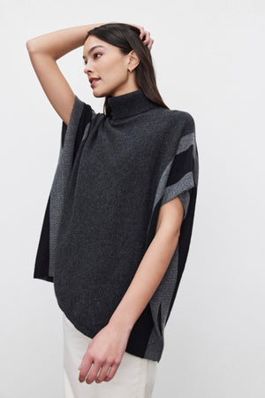 A person with long dark hair wears the REESE CASHMERE TURTLENECK PONCHO by Velvet by Graham & Spencer, which features a dark gray color with light gray accents. They have their left arm raised, creating a relaxed silhouette as they look to the left.