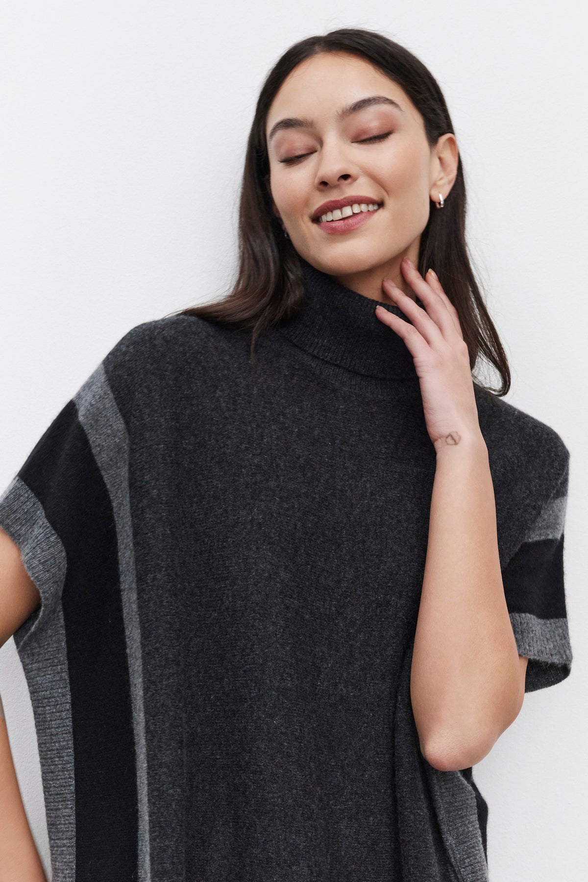   A woman with closed eyes, smiling, while wearing the REESE CASHMERE TURTLENECK PONCHO from Velvet by Graham & Spencer, which features a dark grey color with lighter grey stripe accents and a relaxed silhouette. 