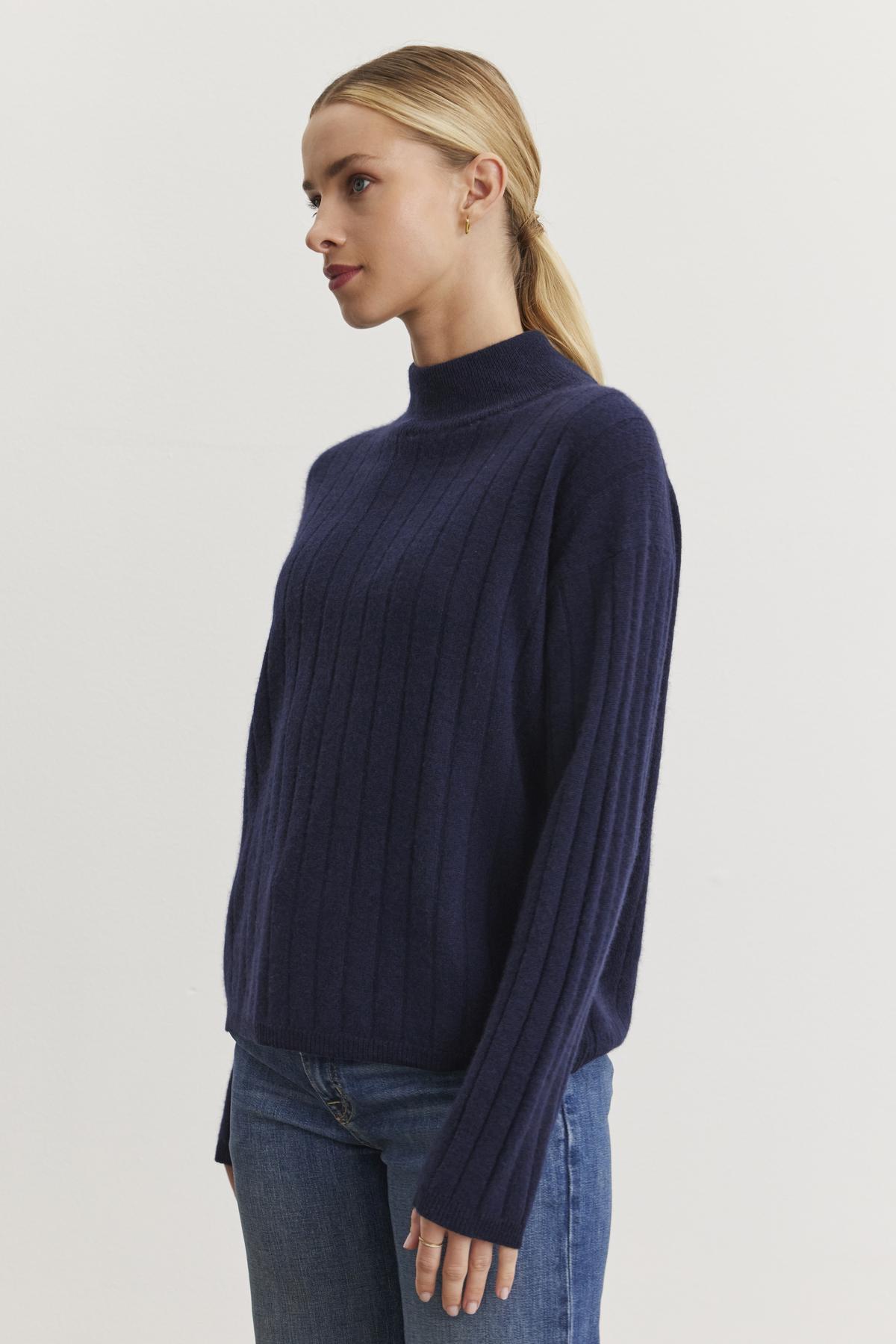   A person with blonde hair, dressed in the luxurious SIMONE CASHMERE MOCK NECK SWEATER by Velvet by Graham & Spencer, which showcases a dark blue hue and ribbed stitch detailing, pairs it with jeans and stands against a plain light background, facing left. 