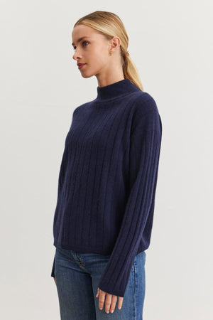 A person with blonde hair, dressed in the luxurious SIMONE CASHMERE MOCK NECK SWEATER by Velvet by Graham & Spencer, which showcases a dark blue hue and ribbed stitch detailing, pairs it with jeans and stands against a plain light background, facing left.