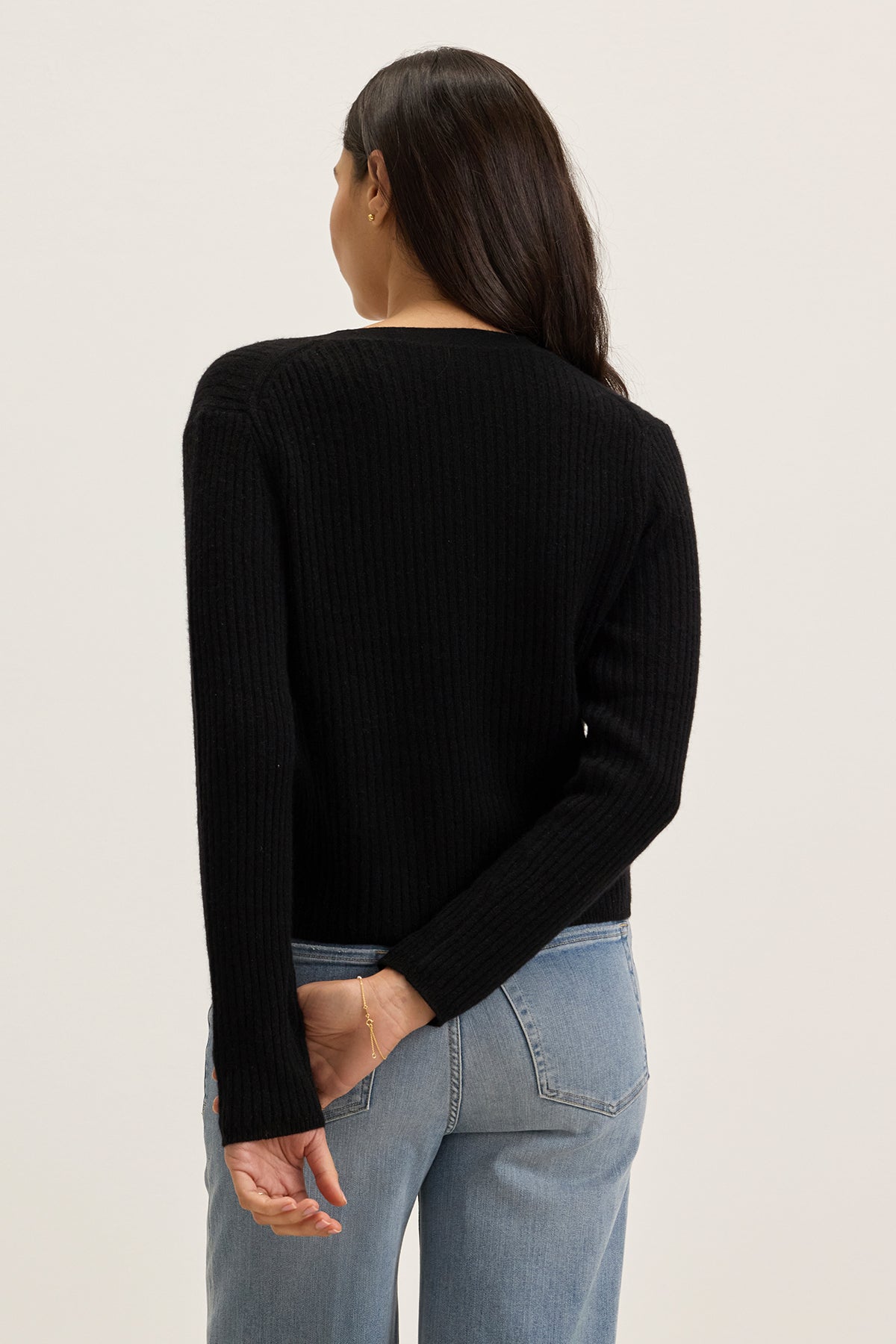 A person with long dark hair stands facing away, wearing the WES CASHMERE CARDIGAN by Velvet by Graham & Spencer, featuring a V-neckline and pearl buttons, paired with light blue jeans and hands crossed behind their back.-39333166874817