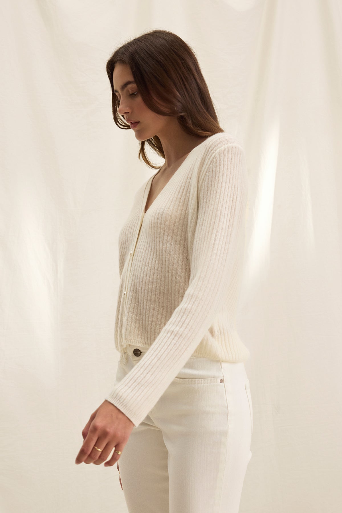 A woman with long brown hair models the Velvet by Graham & Spencer WES CASHMERE CARDIGAN—a cream V-neck style with pearl buttons—paired with white pants, standing against a light, textured background.-39333168775361