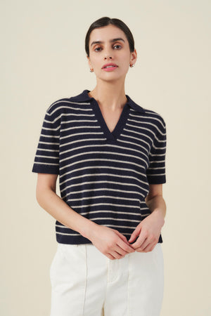 A woman stands against a plain background wearing the Velvet by Graham & Spencer YAEL CASHMERE SWEATER POLO in navy blue and white, featuring a V-neck and striped detailing, paired with white pants for a timeless look.