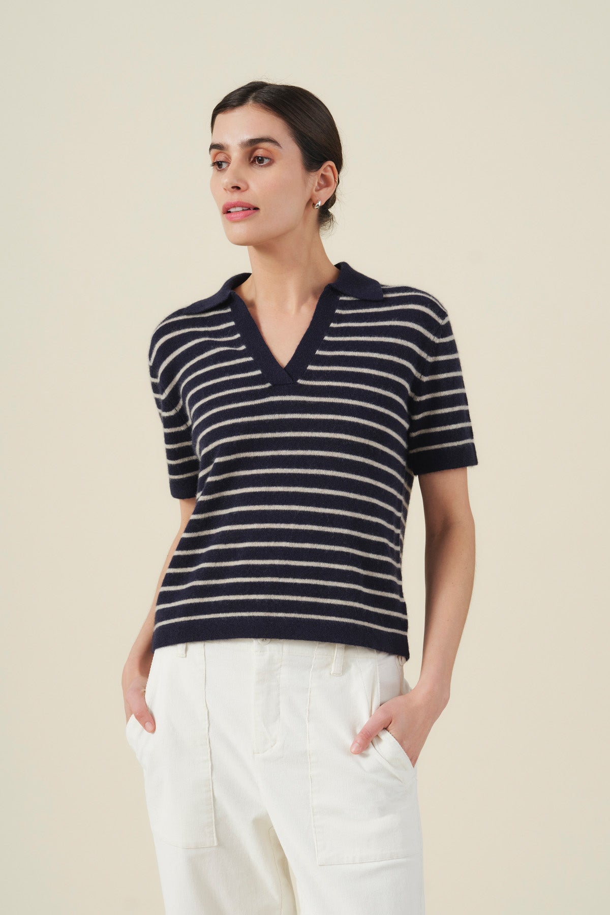 A woman wearing the Velvet by Graham & Spencer YAEL Cashmere Sweater Polo—a navy and white striped short-sleeve knit top with a V-neck collar and refined striped detailing—paired with high-waisted white pants, stands against a plain beige background.