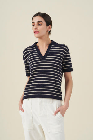 A woman wearing the Velvet by Graham & Spencer YAEL Cashmere Sweater Polo—a navy and white striped short-sleeve knit top with a V-neck collar and refined striped detailing—paired with high-waisted white pants, stands against a plain beige background.